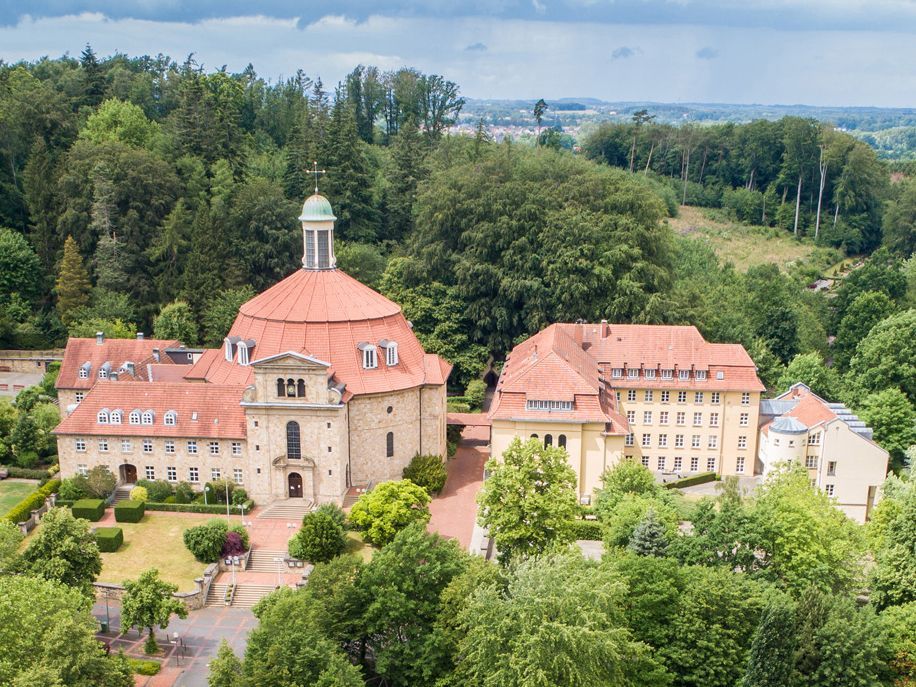 Die Luftaufnahme zeigt den gesamten Gebäudekomplex von Haus Ohrbeck sowie den Konvent der Franziskaner und die Pfarr- und Klosterkirche St. Antonius. Das Ensemble ist eingefasst in das Grün von Bäumen, Sträuchern und des Waldgebietes "Boberg".
